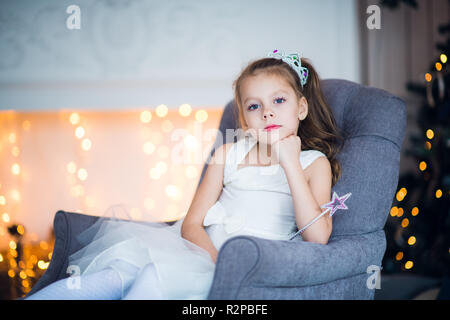 Happy happy little girl wearing crown excité à la veille de Noël, assis sous l'arbre lumineux décoré. Carte de vœux ou couvrir, avec copie espace horizontal. Banque D'Images