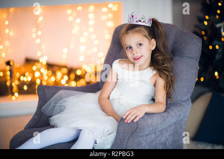 Happy happy little girl wearing crown excité à la veille de Noël, assis sous l'arbre lumineux décoré. Carte de vœux ou couvrir, avec copie espace horizontal. Banque D'Images