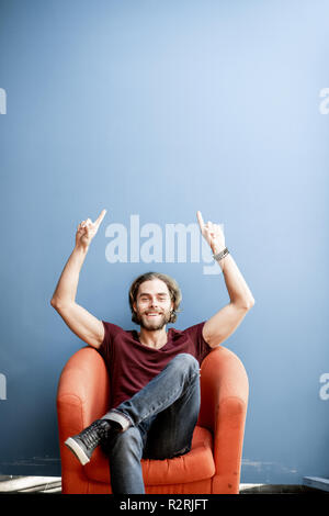 Portrait of a young caucasian homme barbu aux cheveux longs montrant avec les mains sur l'arrière-plan coloré assis sur la chaise. Image avec copie espace Banque D'Images