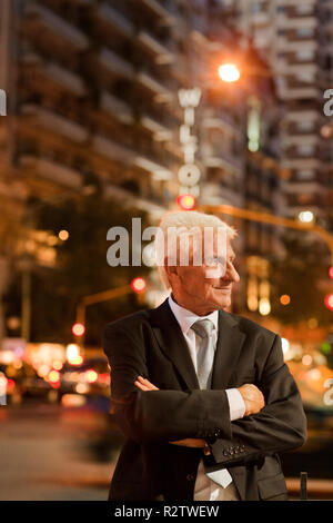 Mature businessman, les bras croisés regarde ailleurs qu'il pose pour un portrait tout en se tenant sur une rue bondée de nuit. Banque D'Images