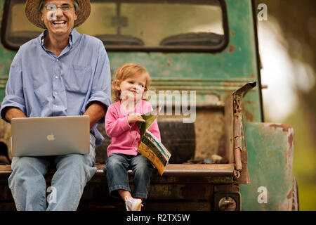 Petite fille à la photo au livre alors que son grand-père utilise son ordinateur portable à l'arrière de sa camionnette. Banque D'Images