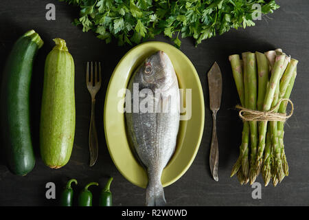 Vue de dessus d'une matière première fraîche dorada poisson dans un plat de vert avec des légumes verts et des couverts sur une table noire Banque D'Images