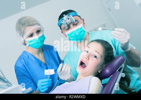 Portrait de peur teen girl avec la bouche ouverte pendant un traitement dentaire avec deux dentistes debout près de présidence Banque D'Images