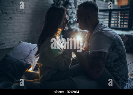 Jeune couple dans l'amour est assis sur le plancher et tenir la main sur fond d'arbre de Noël, ampoules rougeoyant, mur de briques et de fenêtre. Banque D'Images