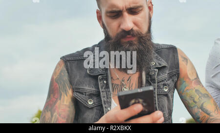 Homme barbu et brutal avec des tatouages ressemble dans le téléphone dans la rue. Banque D'Images