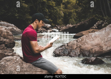 Man relaxing in the woods tout en lisant un livre près d'une rivière dans la nature Banque D'Images