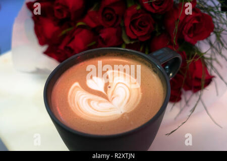 Sur le café crème en forme de coeur avec des roses rouges bouquet sur table Banque D'Images