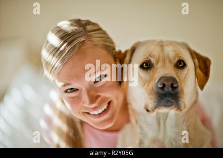 Souriante jeune femme embrasse son chien dans sa chambre ensoleillée. Banque D'Images
