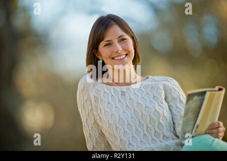 Smiling young woman relaxing with a book in a sunlit jardin. Banque D'Images