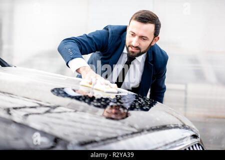 Elegant businessman vêtus de la combinaison lave sa voiture avec une éponge jaune et de la mousse sur un self service lavage de voiture Banque D'Images