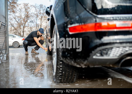 Rondelle professionnel en uniforme noir et les essuyant avec une éponge roue d'une voiture de luxe pendant le processus de lavage à l'extérieur Banque D'Images