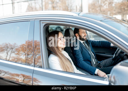 Businessman and woman habillés en costumes de la conduite de voiture de luxe, vue de l'extérieur à travers la fenêtre Banque D'Images