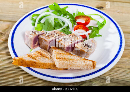 Délicieux thon frit légèrement saupoudré de sésame servi sur une assiette avec du pain grillé, tomates fraîches et vertes. Banque D'Images