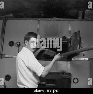 1965, young male Baker en utilisant une palette de bois pour faire ressortir des baguettes de pain d'un grand four commercial ou industriel, England, UK. Banque D'Images