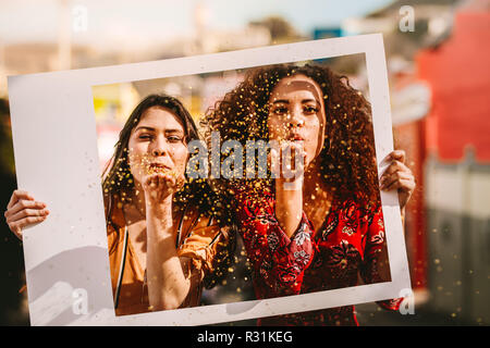 Belles jeunes femmes soufflant une rafale de magie à travers un gros background cadre photo vide à l'extérieur. Amis de sexe féminin attrayant avec soufflage cadre photo Banque D'Images