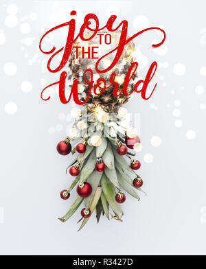 Vue de dessus de boules de Noël rouge sur l'ananas frais isolé sur blanc avec 'joy to the world' inspiration et lumières bokeh Banque D'Images