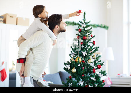 Petite fille assise sur le père en arrière tout en mettant l'étoile décorative rouge en haut de l'arbre de Noël Banque D'Images