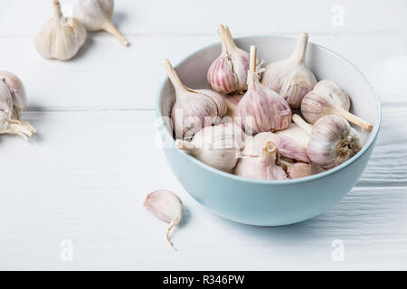 Bol en céramique bleu avec des têtes d'ail et plusieurs gousses pelées sur table de cuisine en bois blanc Banque D'Images