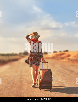 Cute femme en robe d'été et de l'auto-stop avec valise à chapeau aux couleurs de l'automne. Femme marche sur la route avec une assurance. Concept de la liberté Banque D'Images