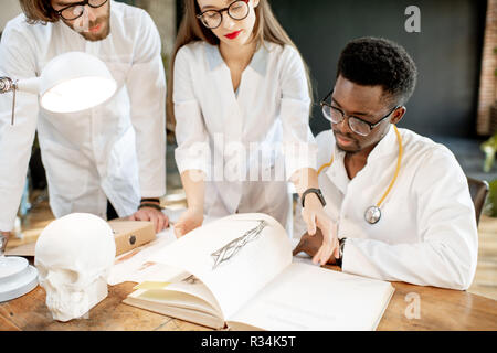 Le groupe d'un jeune multi ethnicité les médecins ou étudiants en médecine en uniforme travaillant avec réserve et des dessins sur la table dans le retro office interior Banque D'Images