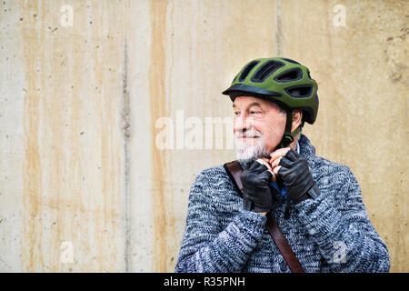 Senior homme debout à l'extérieur contre un mur en béton, en mettant sur casque de vélo. Banque D'Images