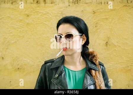 Jeune femme moderne à la mode des vêtements de cigarette dans l'avant du mur jaune. Banque D'Images