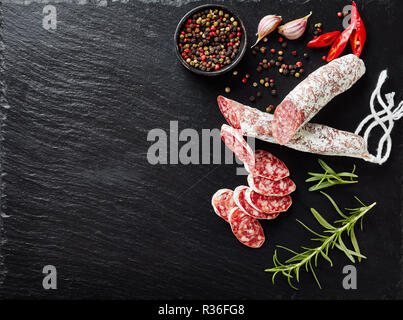 Saucisson Fuet coupé en tranches sur une plaque en ardoise noire avec des grains de poivre, des tranches de piment et du romarin frais, vue de dessus, close-up Banque D'Images