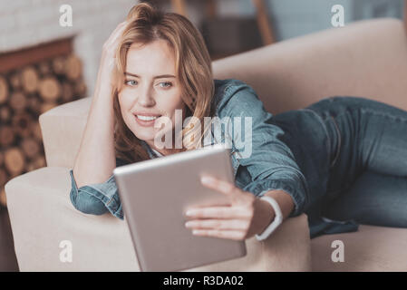 Confort agréable. Jolie femme magnétique couchée sur le canapé tout en prenant vos autoportraits sur sa tablet Banque D'Images