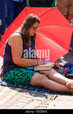 Vue latérale des adultes de grande taille femme assise sur le pavé en plein soleil, lire un livre avec un grand parasol rouge, parapluie, derrière elle. Banque D'Images