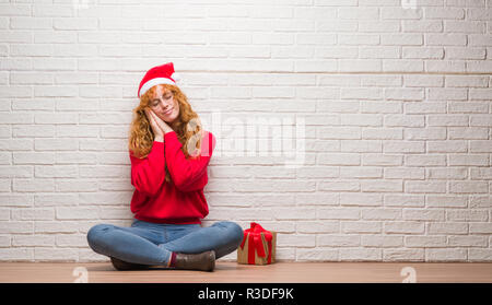 Jeune rousse femme assise sur un mur de briques wearing christmas hat sleeping fatigué de rêver et posant avec les mains tout en souriant avec les yeux fermé Banque D'Images