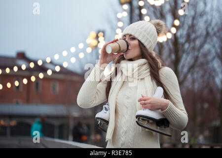 Happy young woman in chandail tricoté et hat va le patinage et boit du café. Jeune brunette avec patins sur son épaule, Parc de la ville en hiver, la glace Banque D'Images