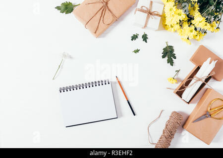 Vue supérieure, ouvrez le bloc-notes avec un crayon blanc sur le tableau blanc télévision décoratif près de jeter l'arrangement de fleurs jaunes, des cadeaux et des bougies, copiez l'espace. Banque D'Images