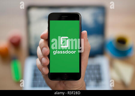 Un homme se penche sur son iPhone qui affiche le logo Glassdoor, tandis qu'assis à son bureau de l'ordinateur (usage éditorial uniquement). Banque D'Images