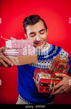 Portrait d'un jeune homme en pull islandais tenant un tas de boîtes-cadeaux sur fond rouge. Noël ou Nouvel An fête concept. Banque D'Images