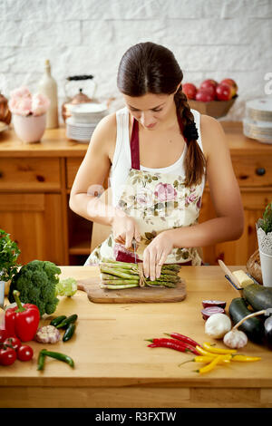 Belle jeune femme brune, l'asperge à trancher dans la cuisine à une table pleine de légumes biologiques Banque D'Images