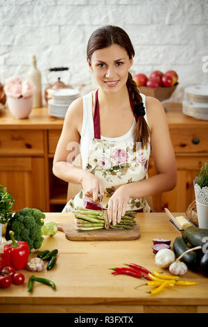 Belle jeune femme brune, l'asperge à trancher dans la cuisine à une table pleine de légumes biologiques Banque D'Images