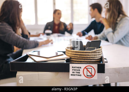 Les téléphones portables, ordinateurs portables et tablettes numériques dans un bac sur table de réunion avec des gens assis à l'arrière et discuter de travail. Réunion sans techno Banque D'Images
