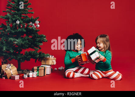 Petites filles, assis près d'un arbre de Noël à jouer avec leurs coffrets cadeaux. Les enfants l'échange de cadeaux de Noël avec un arbre de Noël décoré et une b Banque D'Images