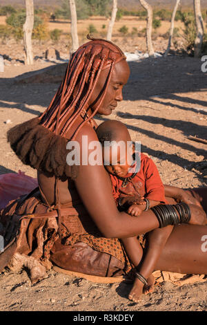 L'Afrique, la Namibie. Femme Himba tient son enfant sur ses genoux, assis sur le sol à l'extérieur de sa hutte dans son village. Banque D'Images