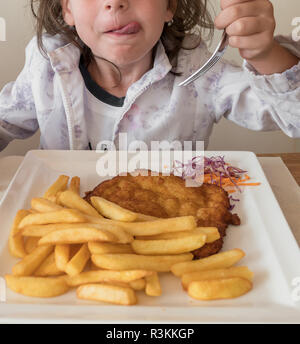 Peu d'italien girl eating viande panées et frites avec une fourchette, dans un restaurant. L'enfant porte manteau de fleurs. Idéal pour les concepts. Banque D'Images