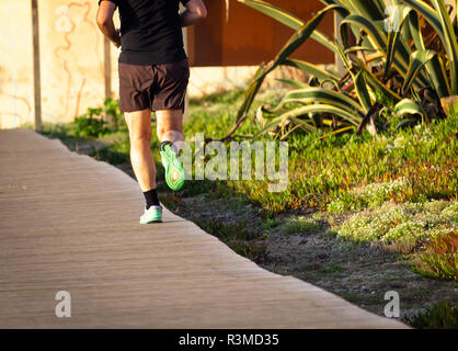 Un homme s'exécute sur un trottoir avec son pied droit vers le haut. Short marron, vert baskets. Vue arrière. Banque D'Images