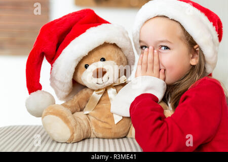 Cute young girl wearing santa hat whispering a secret à son ours en peluche jouet cadeau de Noël. Kid chauds secrets partage avec nounours. Banque D'Images
