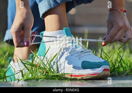 Jeune fille portant des jeans attacher ses lacets de sneakers debout sur l'herbe verte side view close-up photo horizontale, journée ensoleillée Banque D'Images