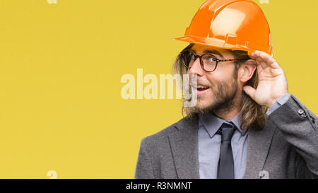 Beau jeune homme architecturaux aux cheveux longs portant un casque de sécurité sur fond isolé smiling with hand sur l'écoute d'une oreille à l'audience ou de rumeur Banque D'Images