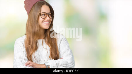 Belle jeune femme brune portant des lunettes et hipster hiver chapeau sur fond isolé à sourire sur le côté avec les bras croisés convaincu Banque D'Images