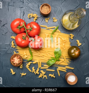 Différents types de pâtes, disposés avec les différents ingrédients, les tomates, les herbes, l'huile et les assaisonnements dans un décanteur sur un fond sombre, rustique sain ve Banque D'Images
