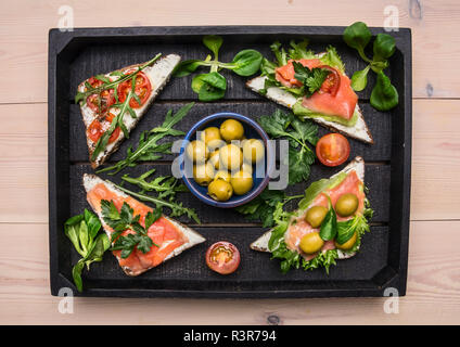 Des sandwichs avec du poisson rouge salé, tomates cerise, roquette, fromage cottage et les herbes dans un bac en bois noir, vue du dessus Banque D'Images