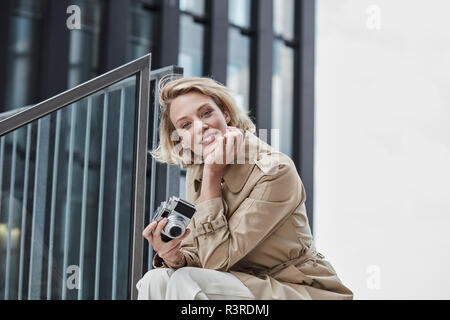 Portrait of smiling blonde woman with digital camera sitting on stairs Banque D'Images