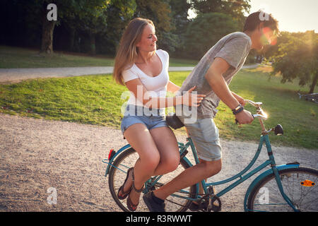 Jeune couple riding bicycle in park, femme assise sur une grille Banque D'Images