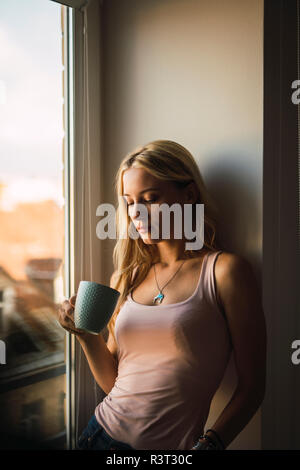 Jeune femme blonde holding coffee mug à la fenêtre Banque D'Images